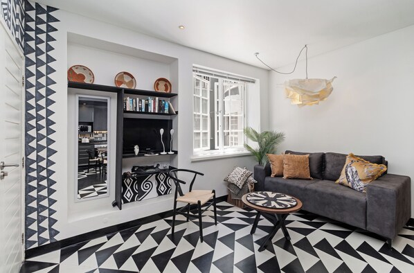 Un salon moderne avec un sol géométrique noir et blanc, un canapé gris agrémenté de coussins à motifs, une petite table ronde et une bibliothèque contenant des objets décoratifs.