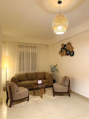 A living room with a beige sofa, two armchairs, a coffee table, a floor lamp, a wall-mounted decorative piece, and a hanging light fixture.