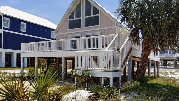 Een tweelaags strandhuis met een balkon, palmbomen en een zandstrand op de achtergrond.