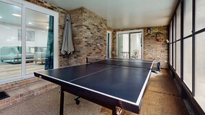 Una mesa de ping pong en una habitación con paredes de ladrillo, ventanas amplias y una puerta corrediza de vidrio que da a un balcón.