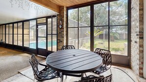 Un patio cubierto con una mesa redonda y sillas, ventanas amplias y una pared de ladrillo.