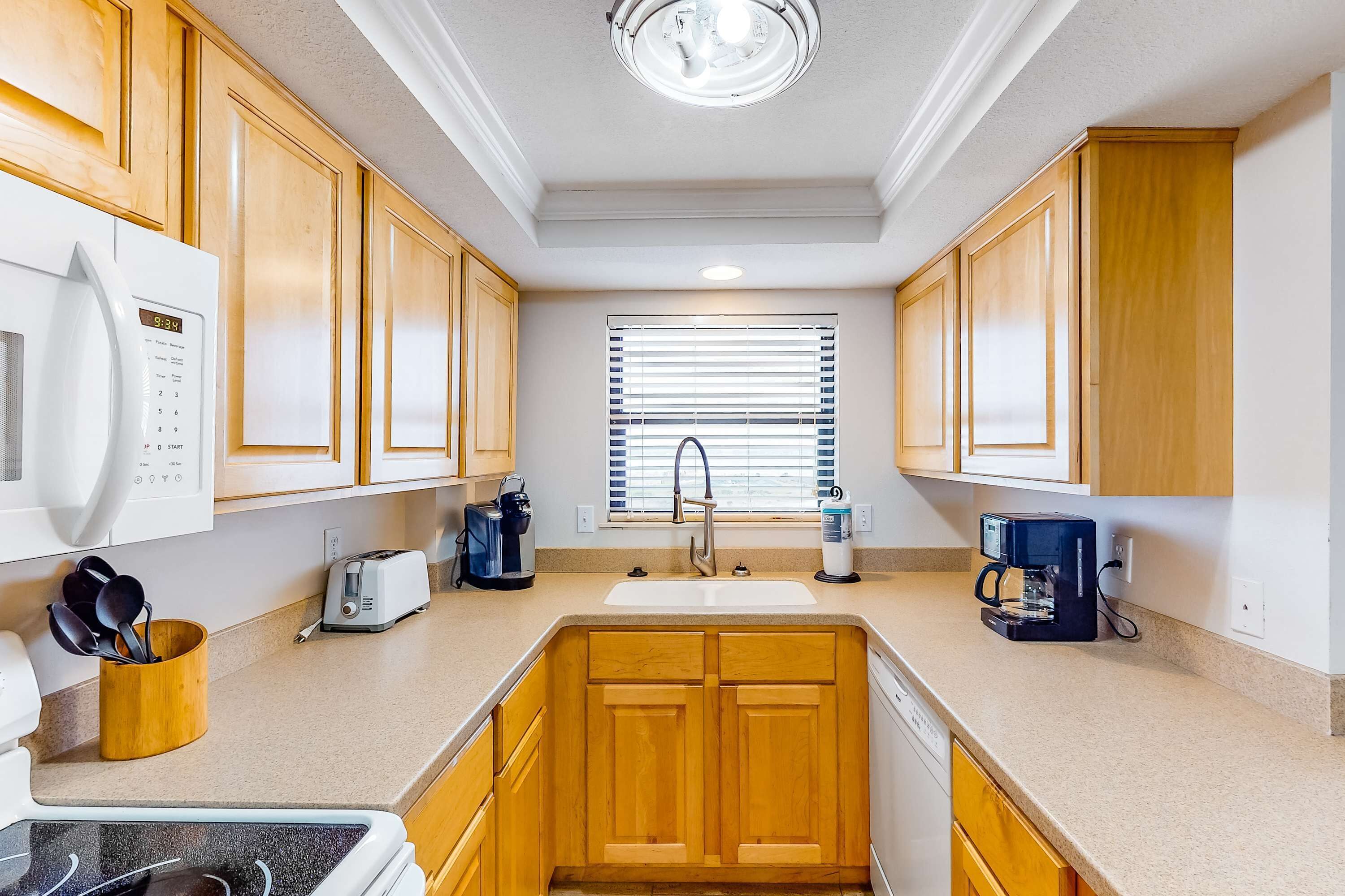 A compact kitchen with wooden cabinets, a microwave, a toaster, a coffee maker, and a window with blinds.