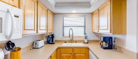 A compact kitchen with wooden cabinets, a microwave, a toaster, a coffee maker, and a window with blinds.