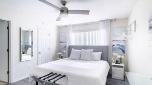 Un dormitorio moderno con una cama grande, un ventilador de techo, una mesita de noche con lámpara y una ventana con cortinas.