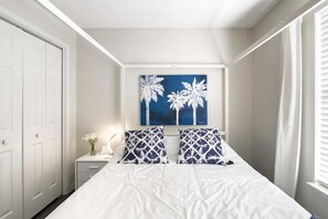 Une chambre à coucher avec un grand lit, deux oreillers, une table de chevet avec une lampe, un tableau représentant des palmiers et une armoire.
