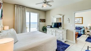 Habitación de hotel con cama, ventilador de techo, televisión, cómoda y vista al océano.