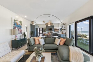 A modern living room with a sectional sofa, a coffee table, and a view of the kitchen and outdoor seating area.