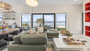 Un salon moderne doté d’une grande fenêtre donnant sur la plage, un coin repas avec une table en bois et une bibliothèque rouge.