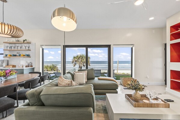 Una sala de estar moderna con un ventanal grande que da a la playa, un comedor con una mesa de madera y una estantería roja.