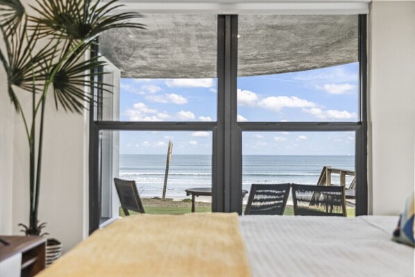 A room with a large window overlooking a beach, a bed, a plant, and a view of the ocean. - Oceanfront Estate | Rooftop Lounge | Game Room (Daytona Beach)