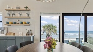 Une cuisine moderne avec vue sur l’océan, comprenant une table à manger en bois, des meubles de rangement gris et de la verrerie décorative exposée sur des étagères.