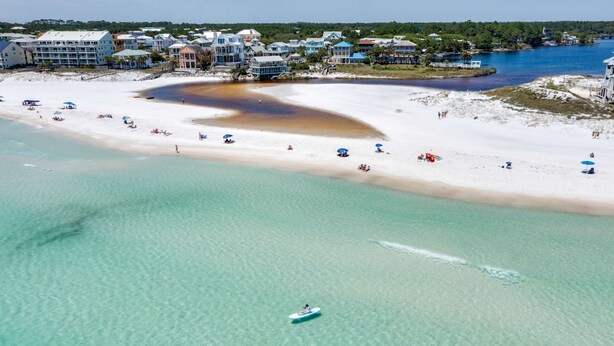 An aerial view of a beach with clear turquoise water, a sandy shore, and several beach umbrellas. - Tree-lined home with deck and AC - 1 block from the beach, Eastern Lake, and 30A (Santa Rosa Beach)
