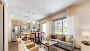 Un salon moderne avec un canapé gris, des fauteuils beige et une table basse. On aperçoit à l’arrière-plan une cuisine avec un coin repas.