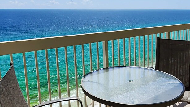 Een balkon met een glazen tafel en stoelen, met uitzicht op een turquoise zee en heldere lucht.
