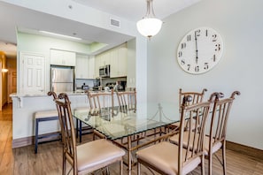 A dining area with a glass table and wooden chairs, a kitchen in the background, and a wall clock. - Sixth Floor, Oceanfront Gulf-Front Condo w/ Shared Pool (Fort Walton Beach)
