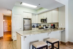 A modern kitchen with white cabinets, a stainless steel refrigerator, a microwave, and a coffee maker. There are two bar stools and a marble countertop. - Sixth Floor, Oceanfront Gulf-Front Condo w/ Shared Pool (Fort Walton Beach)