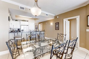 A dining area with a glass table and metal chairs, a kitchen in the background, and beige walls. - Sixth floor Summer Place condo w beach access, shared pool, and more (Fort Walton Beach)