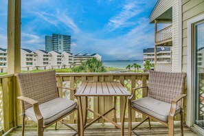 A wooden deck with two wicker chairs and a small table, overlooking a beach and hotel buildings. - Remodeled condo with bay views, pool, fishing pier, & easy beach access (Pensacola Beach)