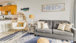 Ett vardagsrum med en grå soffa, en fåtölj i rotting, ett litet sidobord med en lampa och en skylt med texten ”BEACH” på väggen.