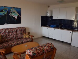 Un espacio habitable compacto con sofá, una mesita redonda y una kitchenette con armarios blancos y salpicadero azul.
