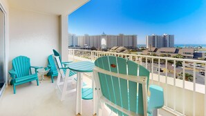 Un balcone con poltrone Adirondack color turchese e un tavolino, con vista su un paesaggio urbano caratterizzato da grattacieli e un cielo azzurro limpido.