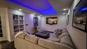 Un soggiorno con un divano color beige, un tavolino da caffè e una TV fissata al muro. Si intravedono anche una libreria e una porta sullo sfondo.