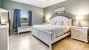 A bedroom with a bed, nightstands, a dresser, a window with curtains, a ceiling fan, and a painting on the wall. - Cozy coastal condo with pool, fast WiFi, central AC, & easy beach access (Pensacola Beach)