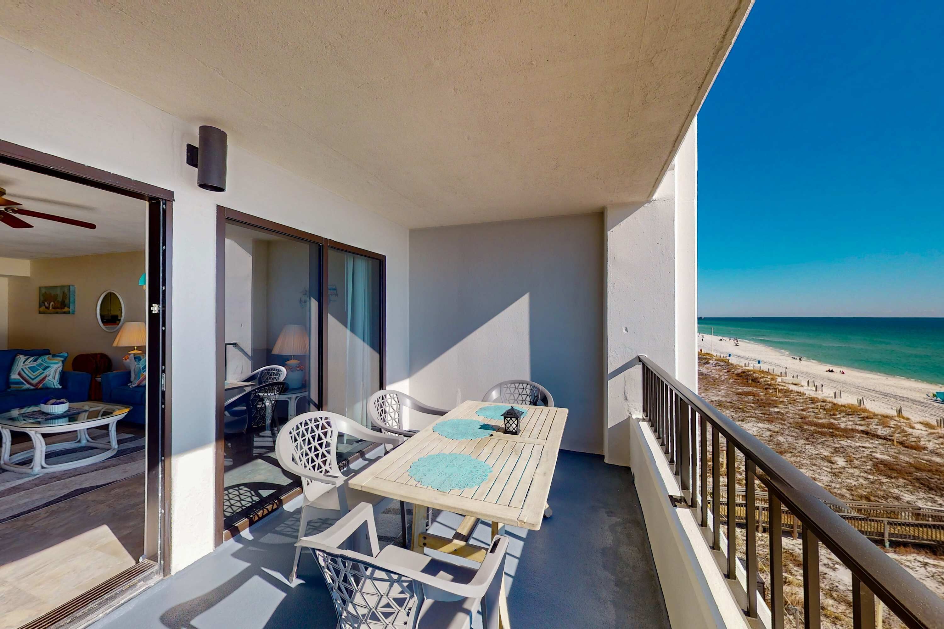 Un balcon avec une table et des chaises, une vue sur la plage et un aperçu de l’intérieur, où l’on aperçoit un canapé et une lampe.