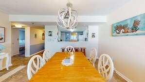Comedor con mesa de madera, sillas blancas, una lámpara de araña y un cuadro de conchas marinas.