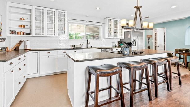 Een moderne keuken met witte kastjes, een groot schiereiland met barstoelen en een hanglamp.