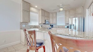 A modern kitchen with light wood cabinets, a granite countertop, stainless steel appliances, and a ceiling fan.