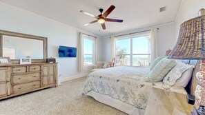 A bedroom with a large bed, a ceiling fan, a dresser, a television, and a view of the ocean through the windows.