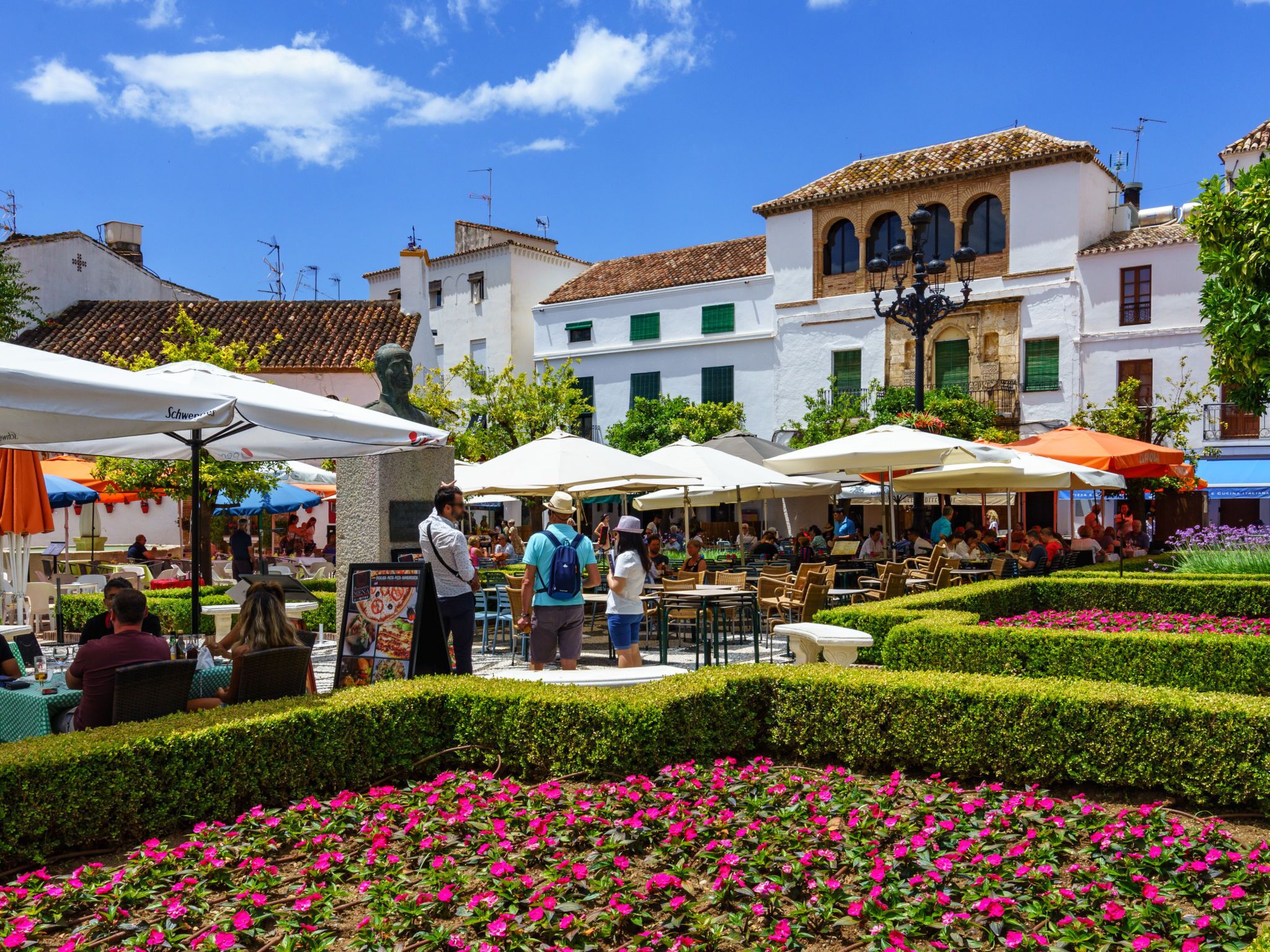 A public square with outdoor seating, a statue, and buildings with traditional architecture.