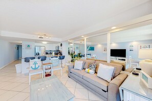 A spacious living area with a sofa, coffee table, and chairs. A kitchen is visible in the background with a dining table and chairs. - 2nd-floor stunning condo with multiple pools, splash pad, & Gulf-front (Destin)