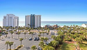 A coastal hotel with a parking lot, palm trees, and a view of the ocean. - Bright beach condo with water views & private balcony - pool, gym & beach (Destin)
