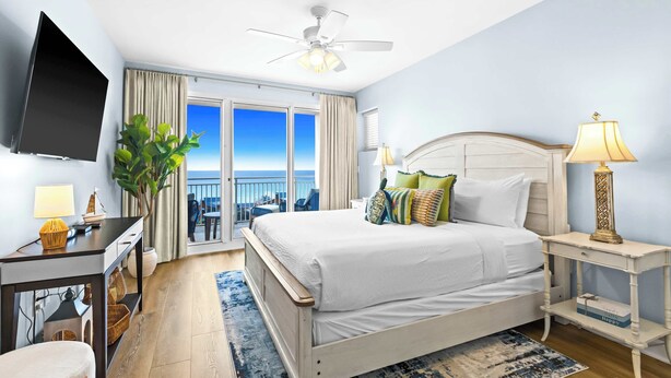 Una camera d'albergo con un letto, una scrivania, una sedia, una TV, un ventilatore a soffitto e vista sull'oceano.