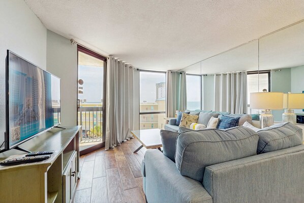 Un salon moderne doté d’un grand téléviseur à écran plat, d’un canapé gris et d’un balcon agrémenté de rideaux.