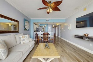 A living room with a sofa, coffee table, and a ceiling fan. There is a dining area with chairs and a table, and a kitchen in the background. - Second Floor Oceanfront Condo with Shared Pools, Ocean View, and Washer&Dryer (Fort Walton Beach)