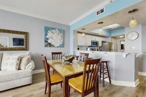 A dining area with a glass table, wooden chairs, and a white sofa. A kitchen is visible in the background with white cabinets and stainless steel appliances. - Second Floor Oceanfront Condo with Shared Pools, Ocean View, and Washer&Dryer (Fort Walton Beach)