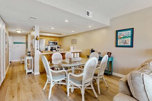 A modern kitchen with white appliances, a dining area with wicker chairs, and a teal side table. - Dreamy Beach Gem W/ Shared Pool, Private Balcony, Water Views & Fast WiFi (Pensacola Beach)