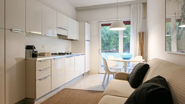 A modern kitchen with white cabinets, a dining area with a white table and chairs, and a view of greenery through the window. - Villa Oliva 8 - Carraro Immobiliare Jesolo (Lido di Jesolo)