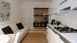 A compact kitchen with a gas stove, white cabinets, and a small dining area with a white table and chair. - Villa Oliva 8 - Carraro Immobiliare Jesolo (Lido di Jesolo)