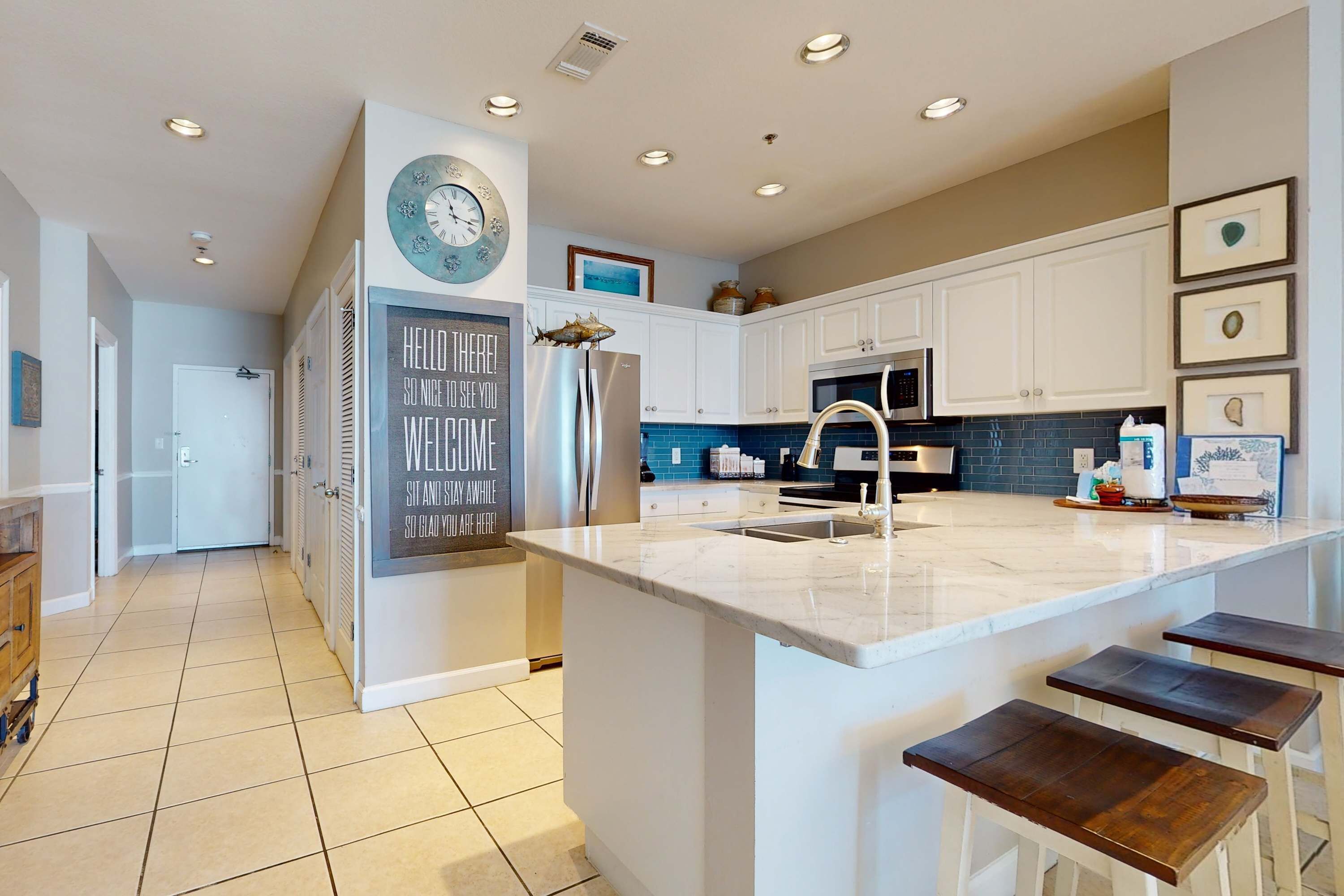 A modern kitchen with white cabinets, a large island with bar stools, a clock, and a welcome sign.