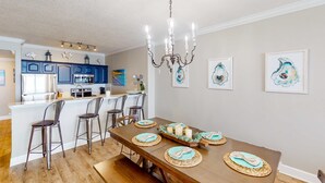 A dining area with a wooden table set for four, surrounded by bar stools. A chandelier hangs above, and there are framed artworks on the wall. - Oceanfront condo with scenic balcony, resort amenities, central AC, & W/D (Panama City Beach)