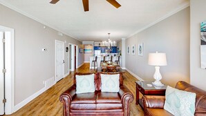 A spacious living room with a brown leather sofa, a dining area in the background, and a ceiling fan. - Oceanfront condo with scenic balcony, resort amenities, central AC, & W/D (Panama City Beach)
