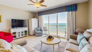 A living room with a television, a red armchair, a grey armchair, a beige sofa, a round coffee table, and a ceiling fan. - Colorful oceanfront condo with Gulf views, on-site pools & hot tub (Panama City Beach)