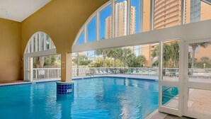 An indoor swimming pool with a view of tall buildings and palm trees outside. - Colorful oceanfront condo with Gulf views, on-site pools & hot tub (Panama City Beach)
