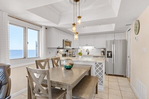 A modern kitchen with white cabinets, a stainless steel refrigerator, and a dining area with a wooden table and chairs. - Oceanfront penthouse with pools, sauna, beach access & breathtaking ocean views (Panama City Beach)