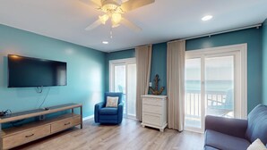 Una sala de estar con televisor, ventilador de techo, un sillón azul, cómoda blanca y vistas al exterior a través de amplios ventanales.