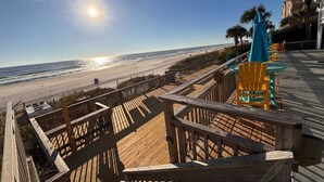 Un paseo marítimo de madera que conduce a una playa, con vista despejada al océano y un edificio con balcón.